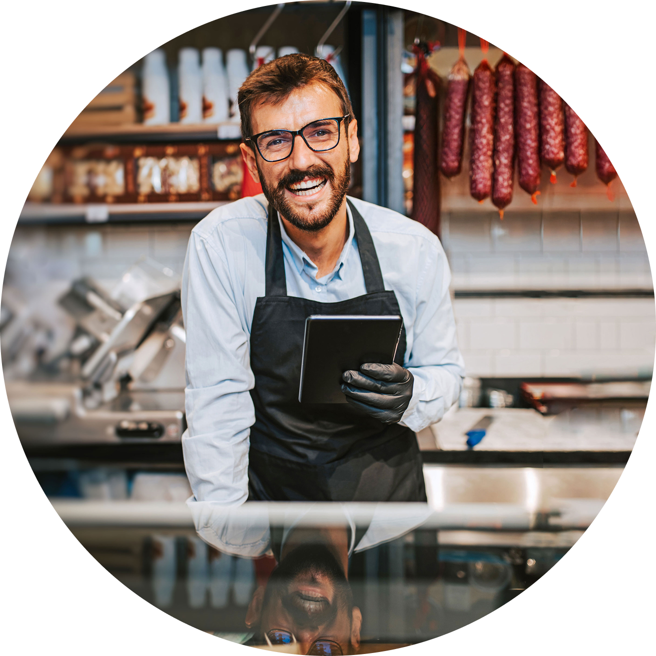 Metzger hinter Verkaufstheke mit Tablet in der Hand. Freundlich lächelnd, im Hintergrund gut präsentierte Wurstwaren.